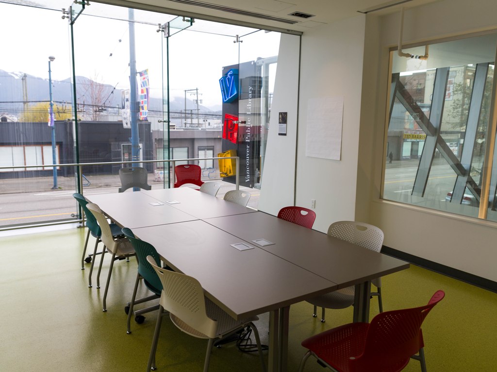 Photograph of Nellie Yip Quong room with tables and chairs set up at the center of a brightly lit room against the backdrop of the city street on the other side of the floor to ceiling window