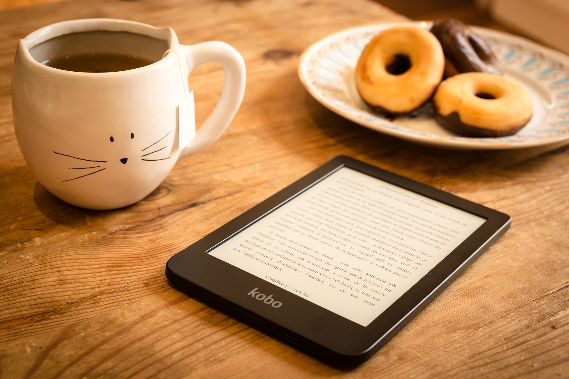 Pictured are an e-reader, cup of tea, and plate of doughnuts on a table. 
