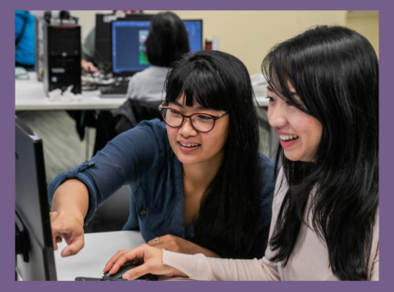 Two women sitting at a computer in the Inspiraton Lab, smiling, while one points at something on the screen. 