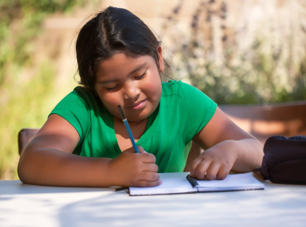 A girl sits at a table, writing in a notebook