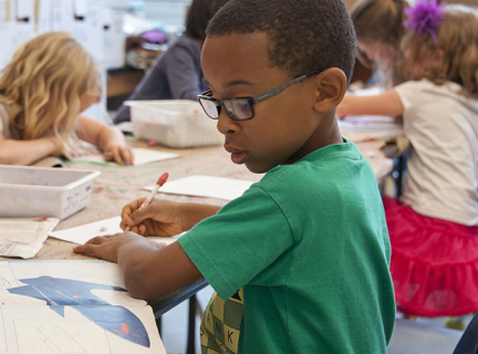 A boy in a green t-shirt and glasses writing with a pencil