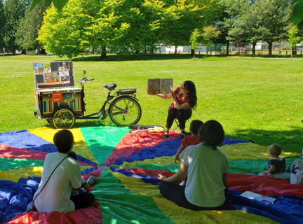 Vancouver Public Library's BiblioBike helps library workers offer storytime outdoors. A Vancouver Public Library worker reads a book to children and their caregivers in a park-like setting.